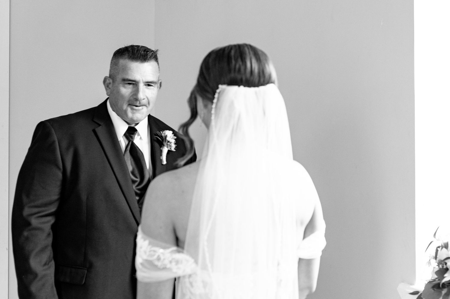 Black and white photo of a groom smiling at his bride in a veil during a wedding first look moment.