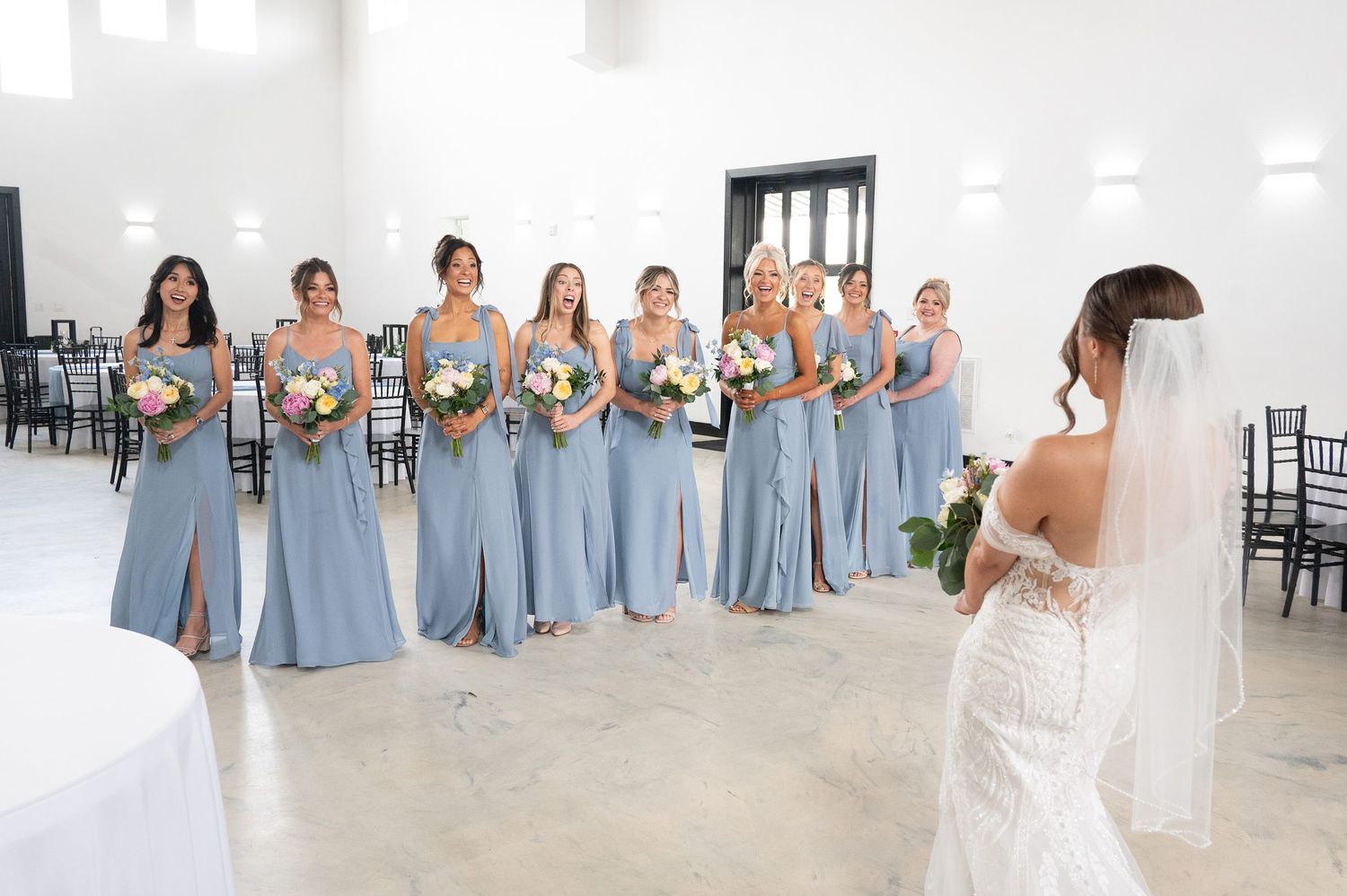 Bride in lace gown faces bridesmaids in dusty blue dresses holding floral bouquets in elegant white wedding venue.