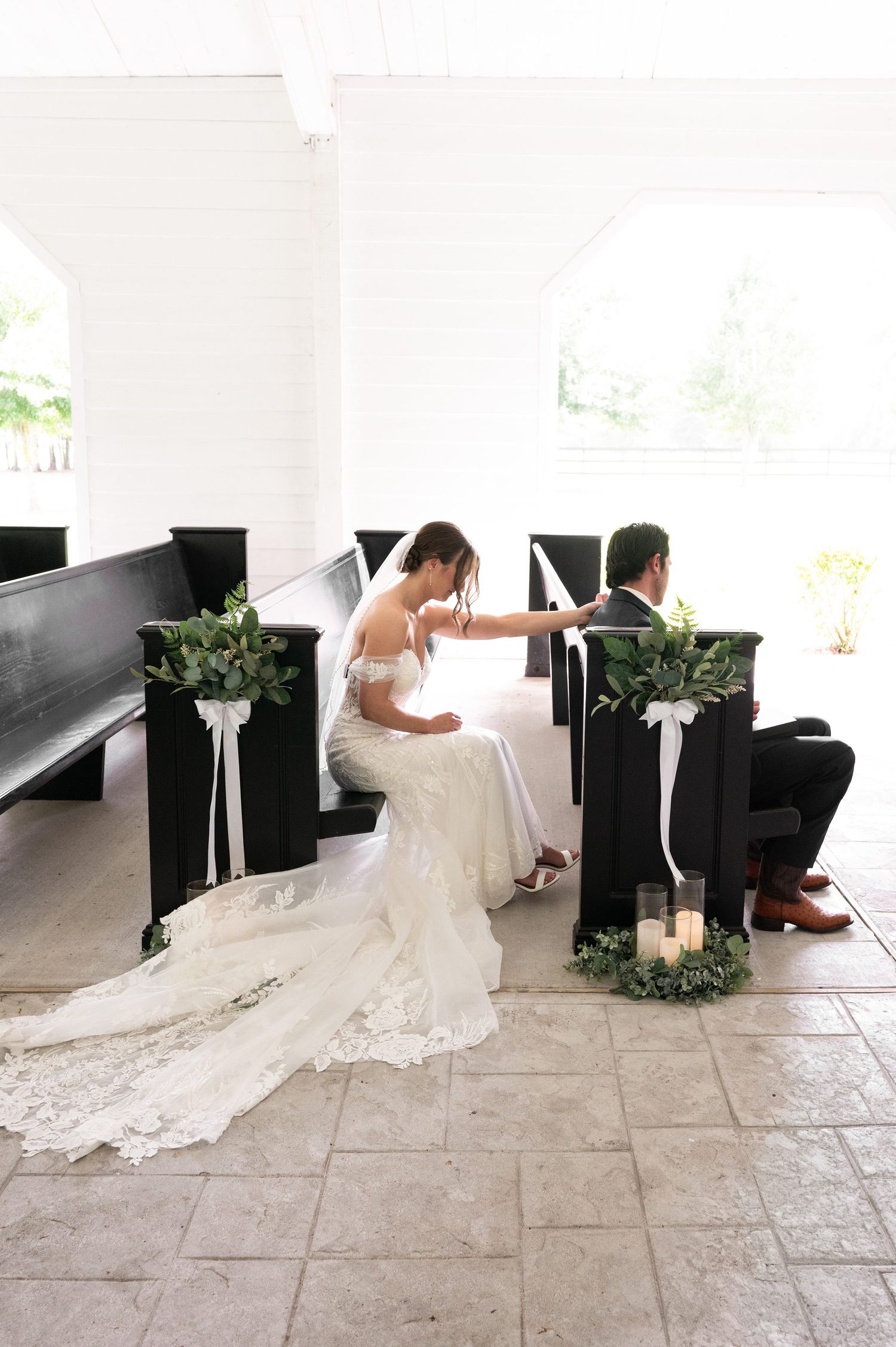 Bride and groom kneeling at church altar during wedding ceremony, surrounded by elegant greenery pew decorations.