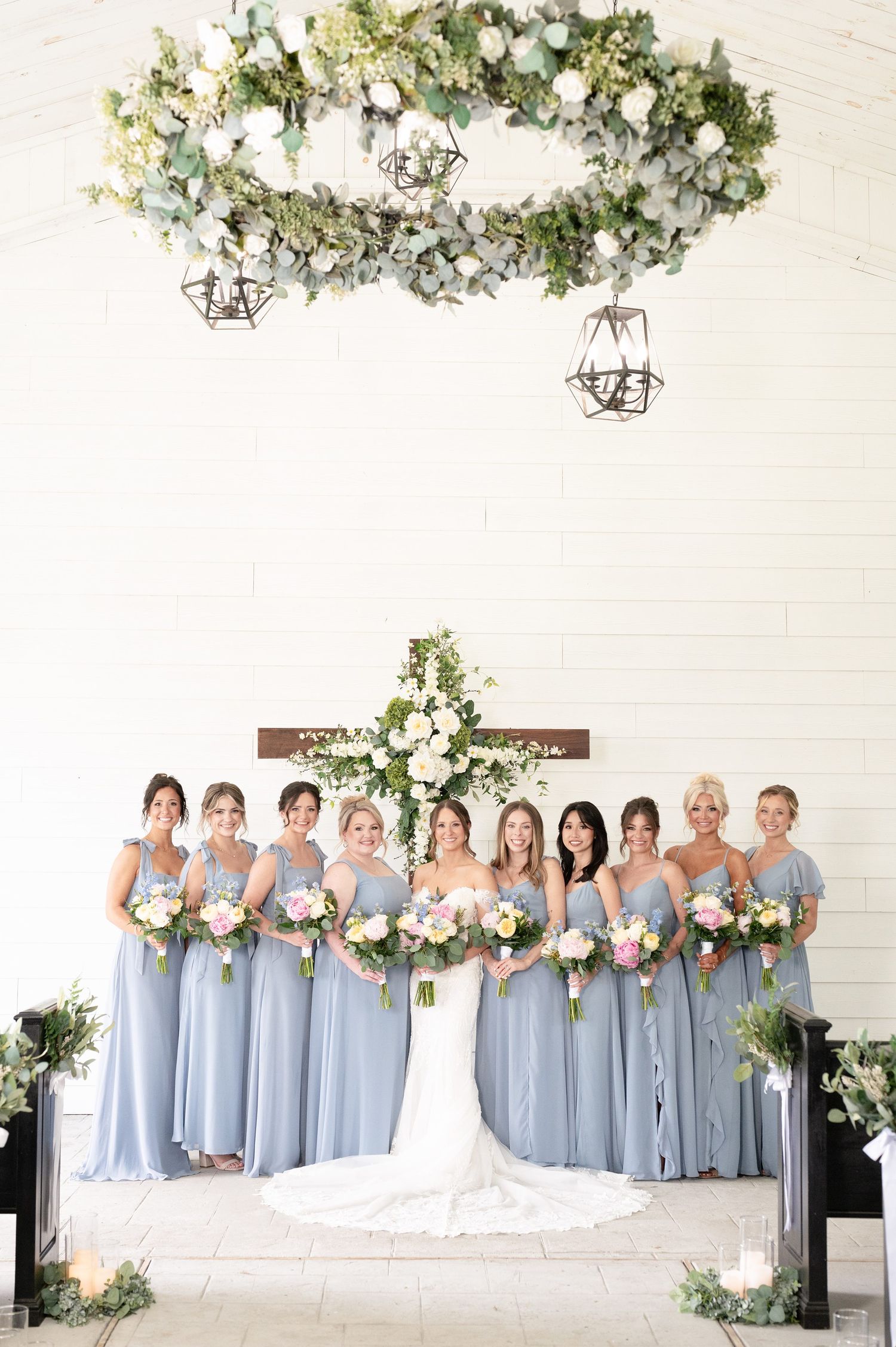 Bride and bridesmaids in dusty blue gowns holding floral bouquets beneath a greenery wreath in a white chapel.