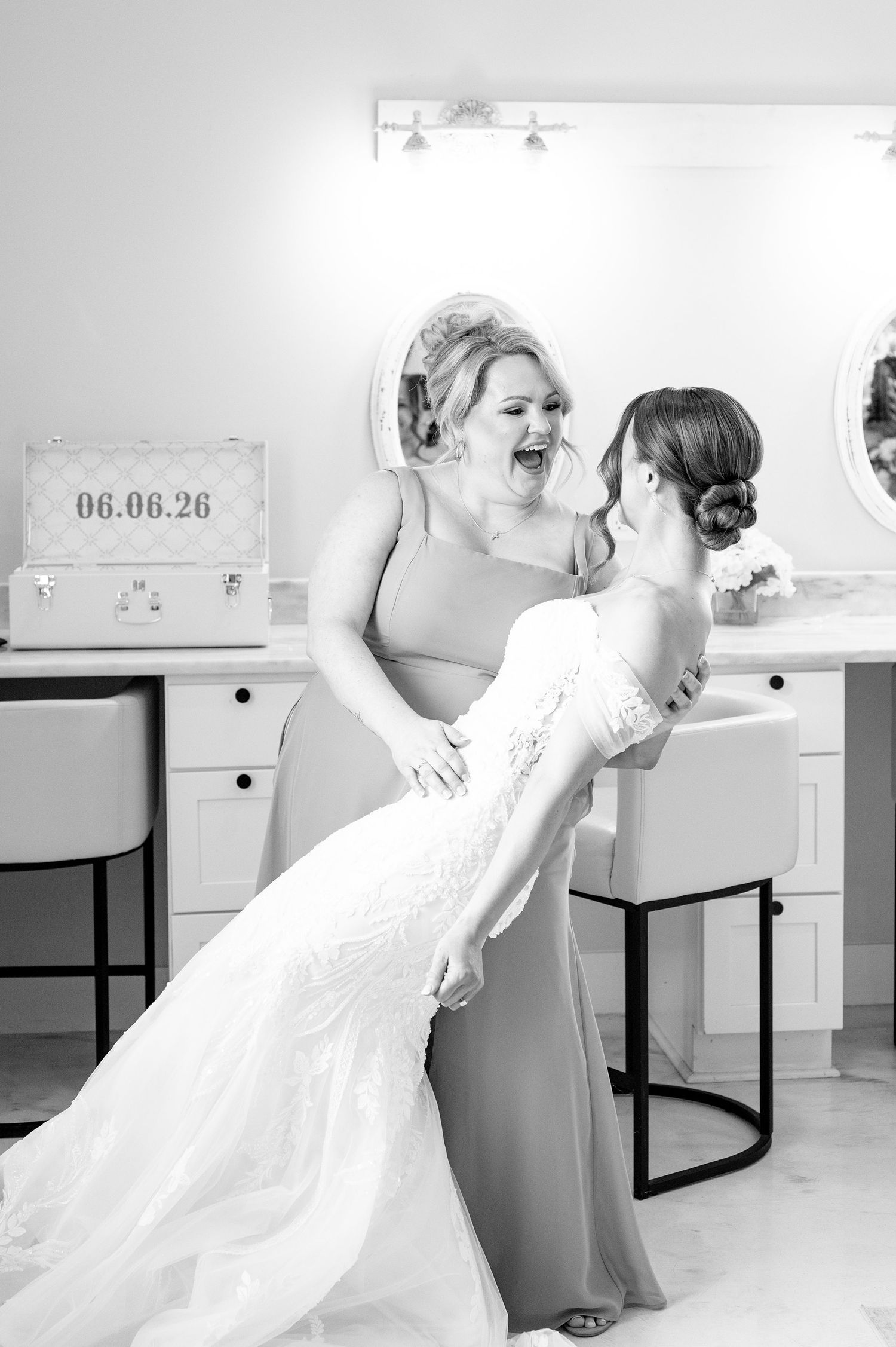 Bride in white lace gown laughing with bridesmaid in dressing room during wedding preparations.