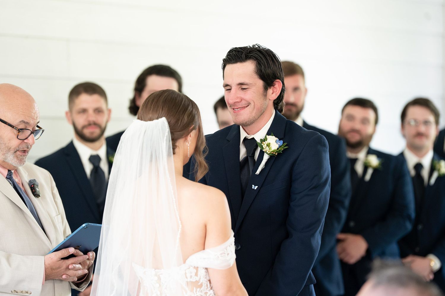Groom smiling at bride during wedding ceremony with groomsmen standing in background at elegant white venue.