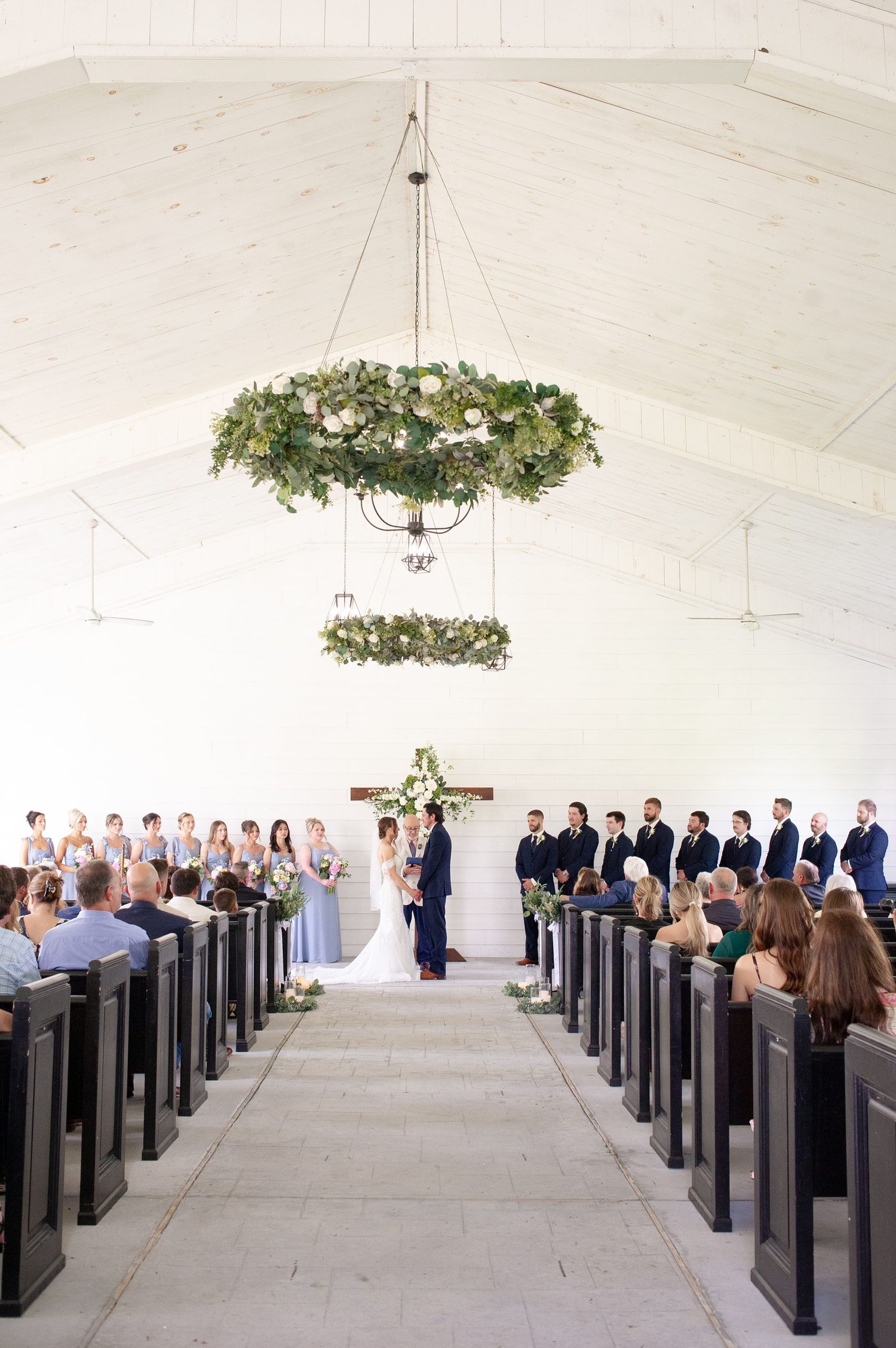 Elegant white chapel wedding ceremony with floral chandeliers, wooden pews, and bridal party at the altar.
