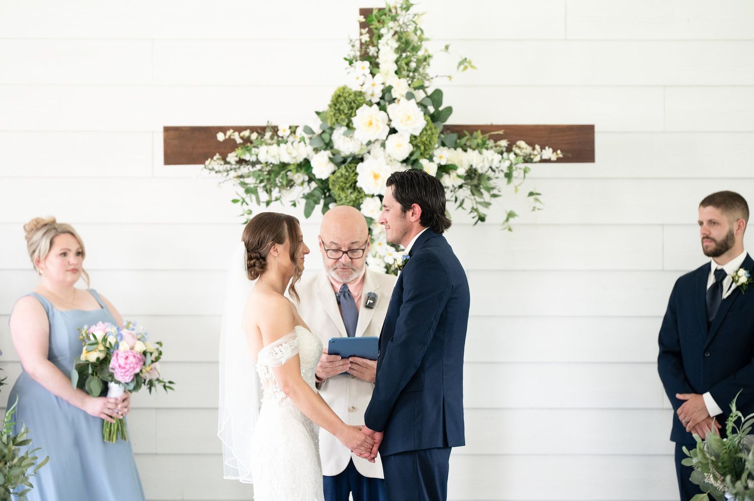 Bride and groom exchange vows at rustic chapel altar adorned with white floral cross arrangement, officiant and wedding party beside them.