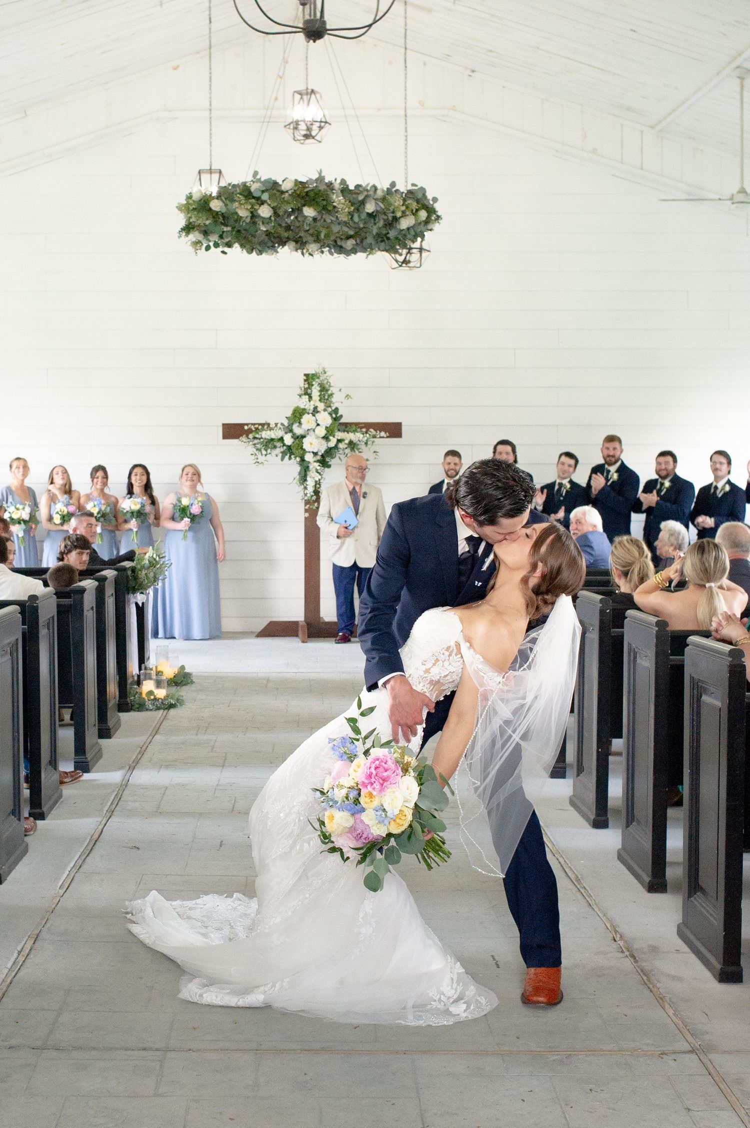 Bride and groom share a romantic dip kiss at the altar in a white chapel with floral chandelier and wedding party.