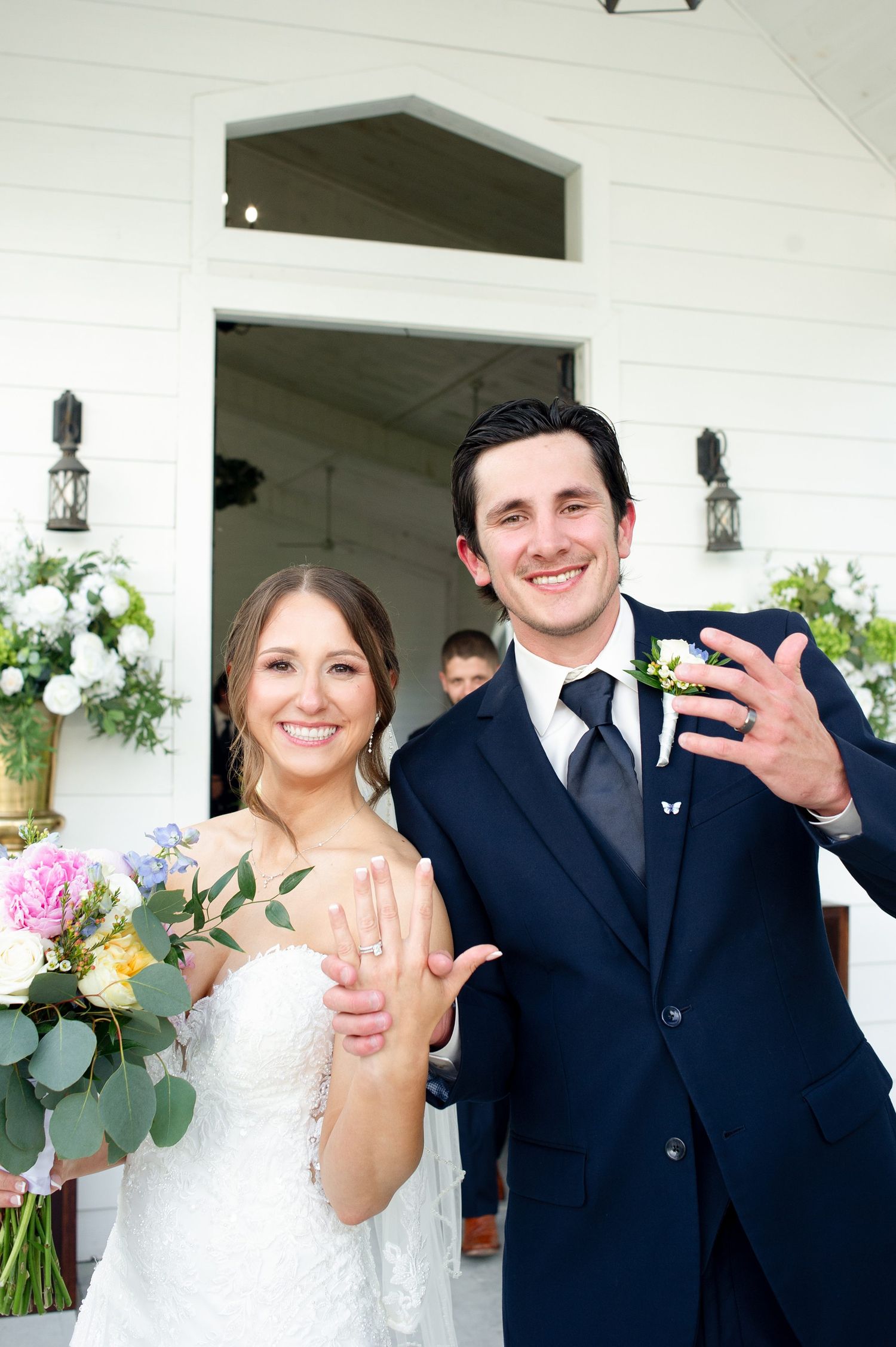 Newlywed couple smiling and showing off wedding rings outside white chapel, bride holding colorful bouquet, groom in navy suit.
