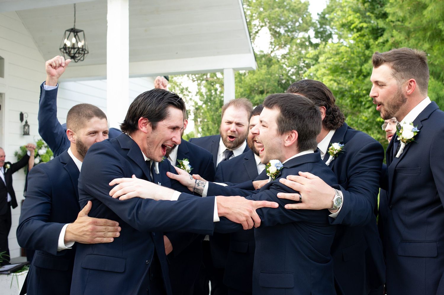 Groomsmen in navy suits celebrate joyfully outside a white venue, laughing and embracing on a wedding day.