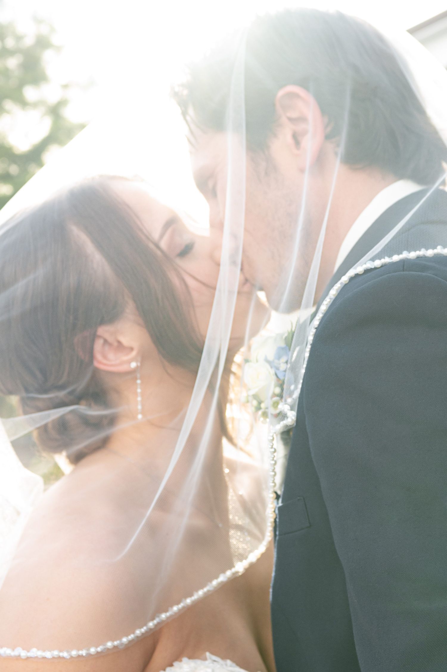 Bride and groom share a romantic kiss under a flowing veil in soft backlit sunlight on their wedding day.