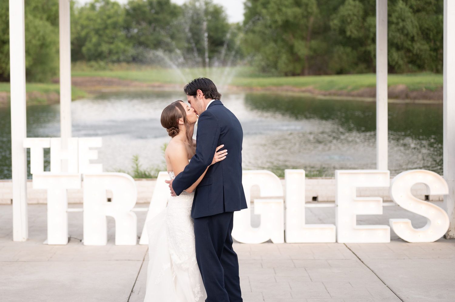 Bride and groom share a kiss in front of large white letter signs near a fountain at an outdoor wedding venue.