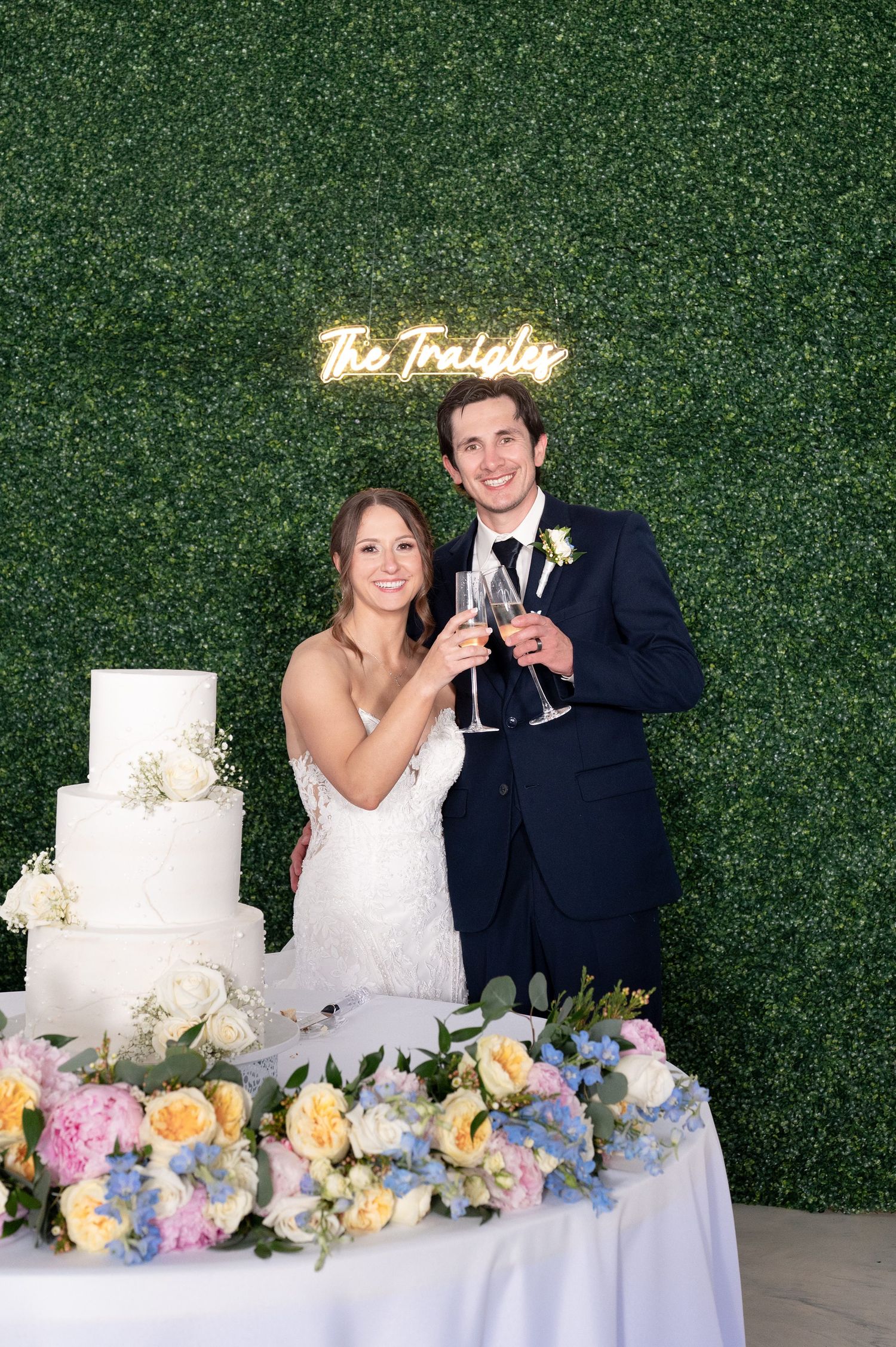 Newlyweds toast with champagne flutes beside a white wedding cake in front of a green hedge wall with neon sign.