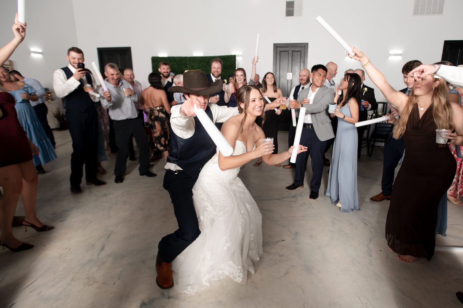 Bride and groom dancing joyfully at wedding reception surrounded by guests holding glow sticks in elegant white venue.