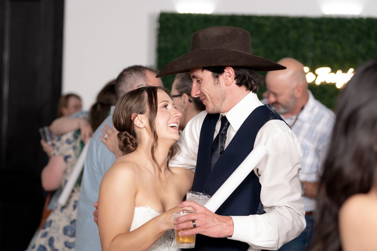 Bride and groom laughing together at wedding reception, groom wearing brown cowboy hat and navy vest.