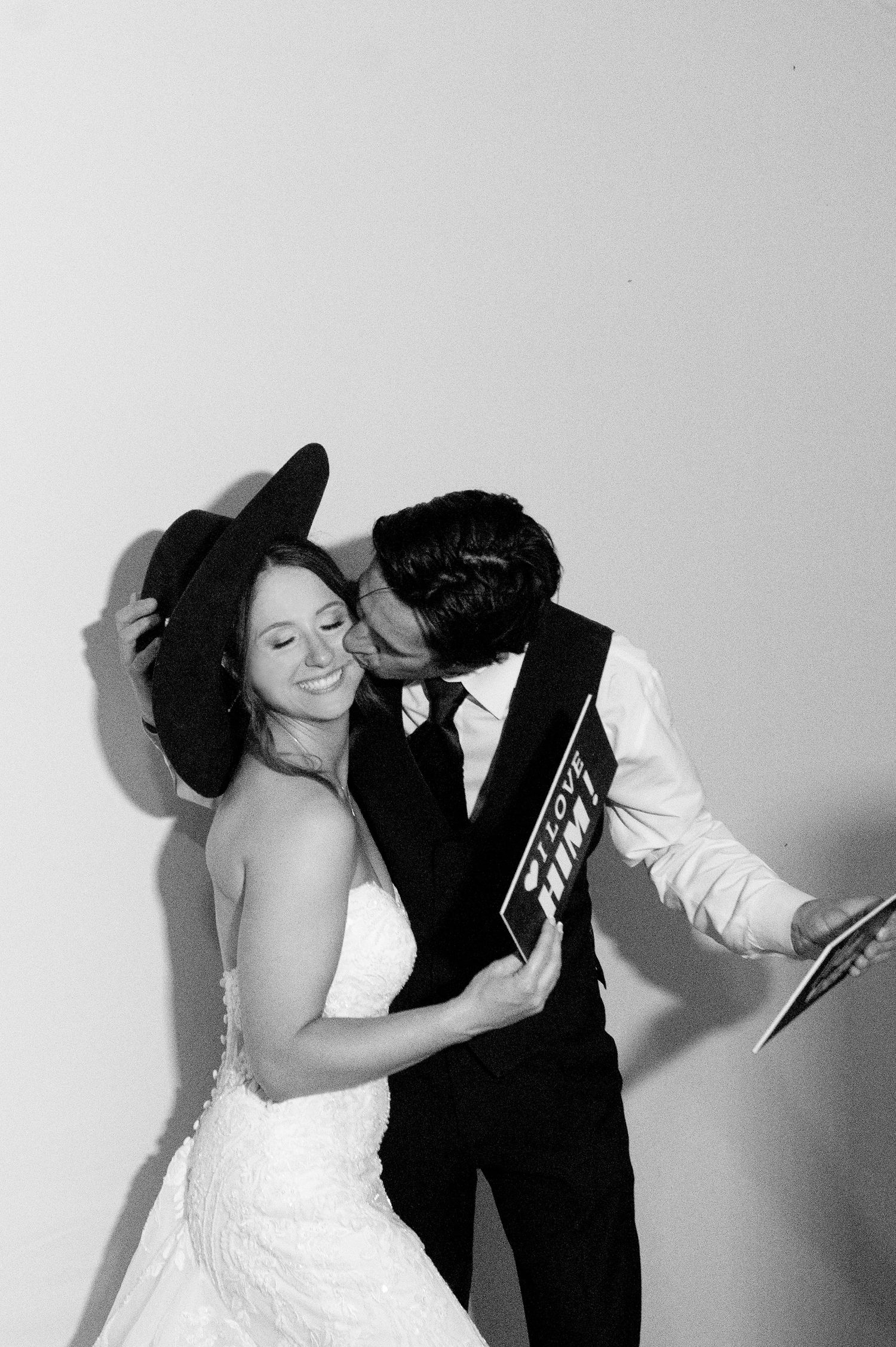 Black and white photo of a smiling bride in white dress being kissed by groom in suit holding a photo booth sign.