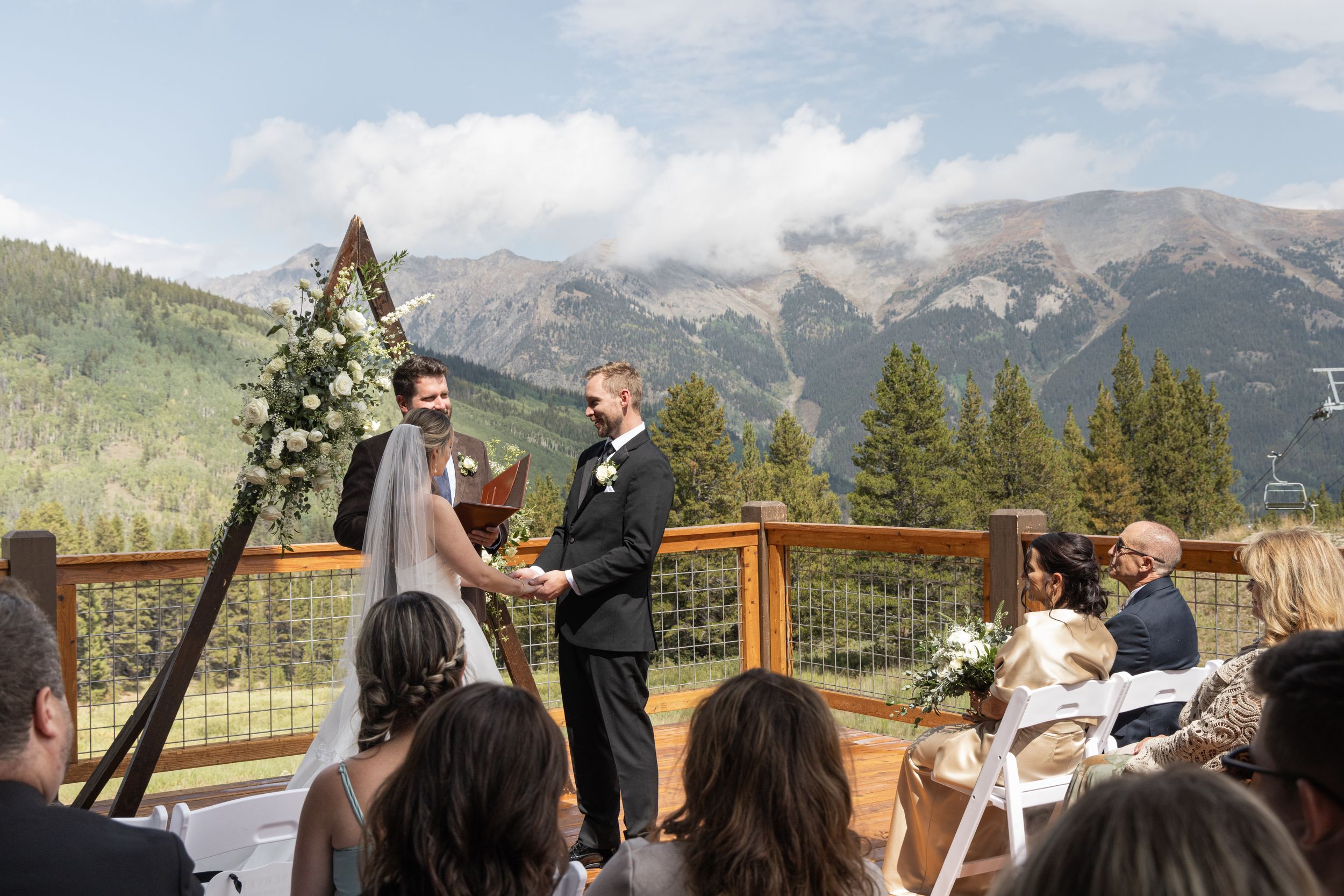 Outdoor mountain wedding ceremony on a deck with couple at floral arch, pine trees, and Rocky Mountain peaks backdrop.