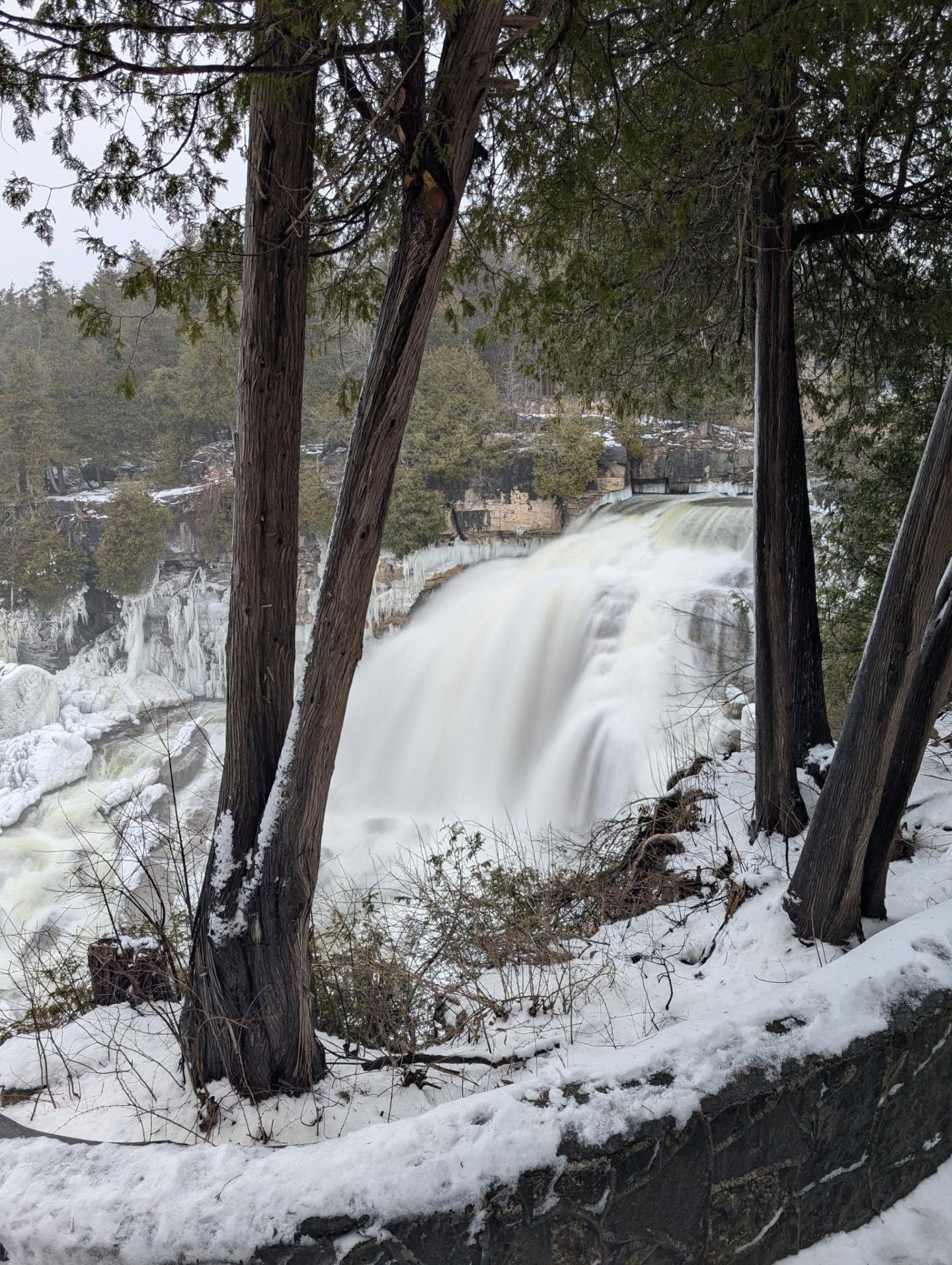 Snow-covered trees frame a powerful waterfall cascading over rocky cliffs in a winter landscape.