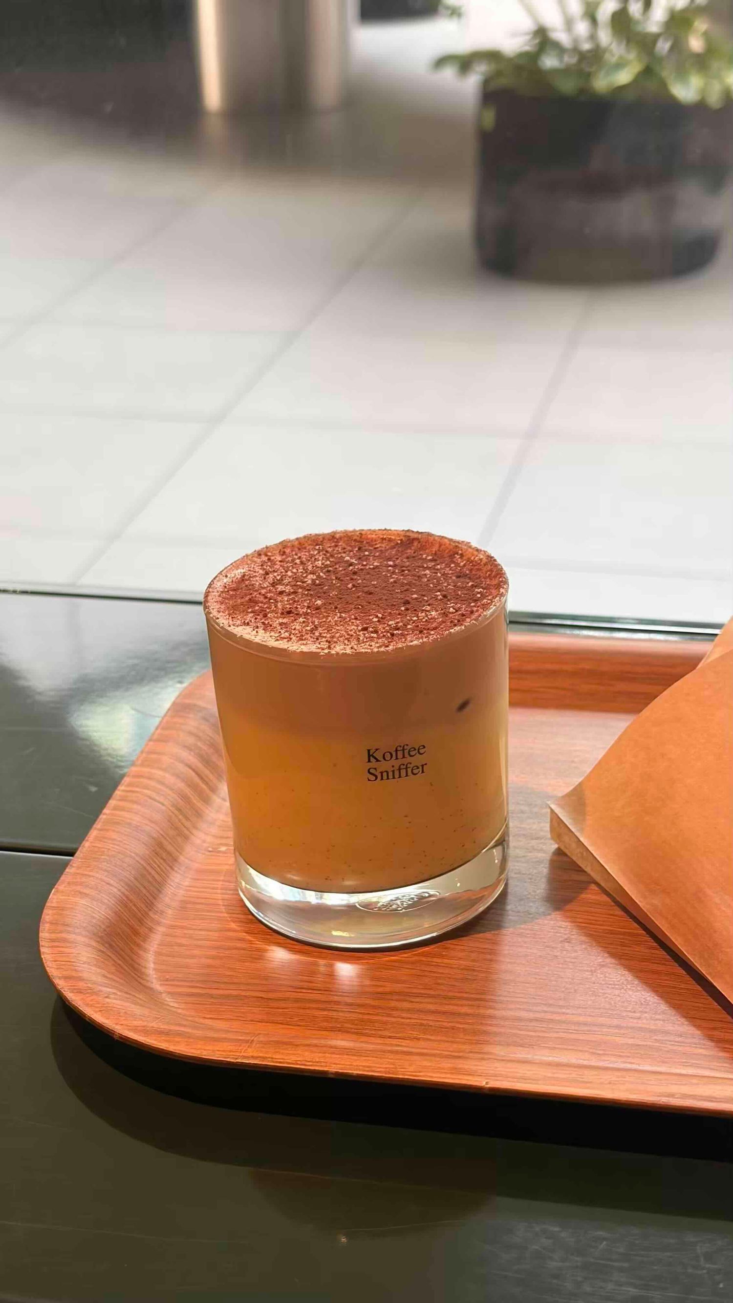 A frothy iced coffee drink dusted with cocoa powder in a glass branded Koffee Sniffer, served on a wooden tray.