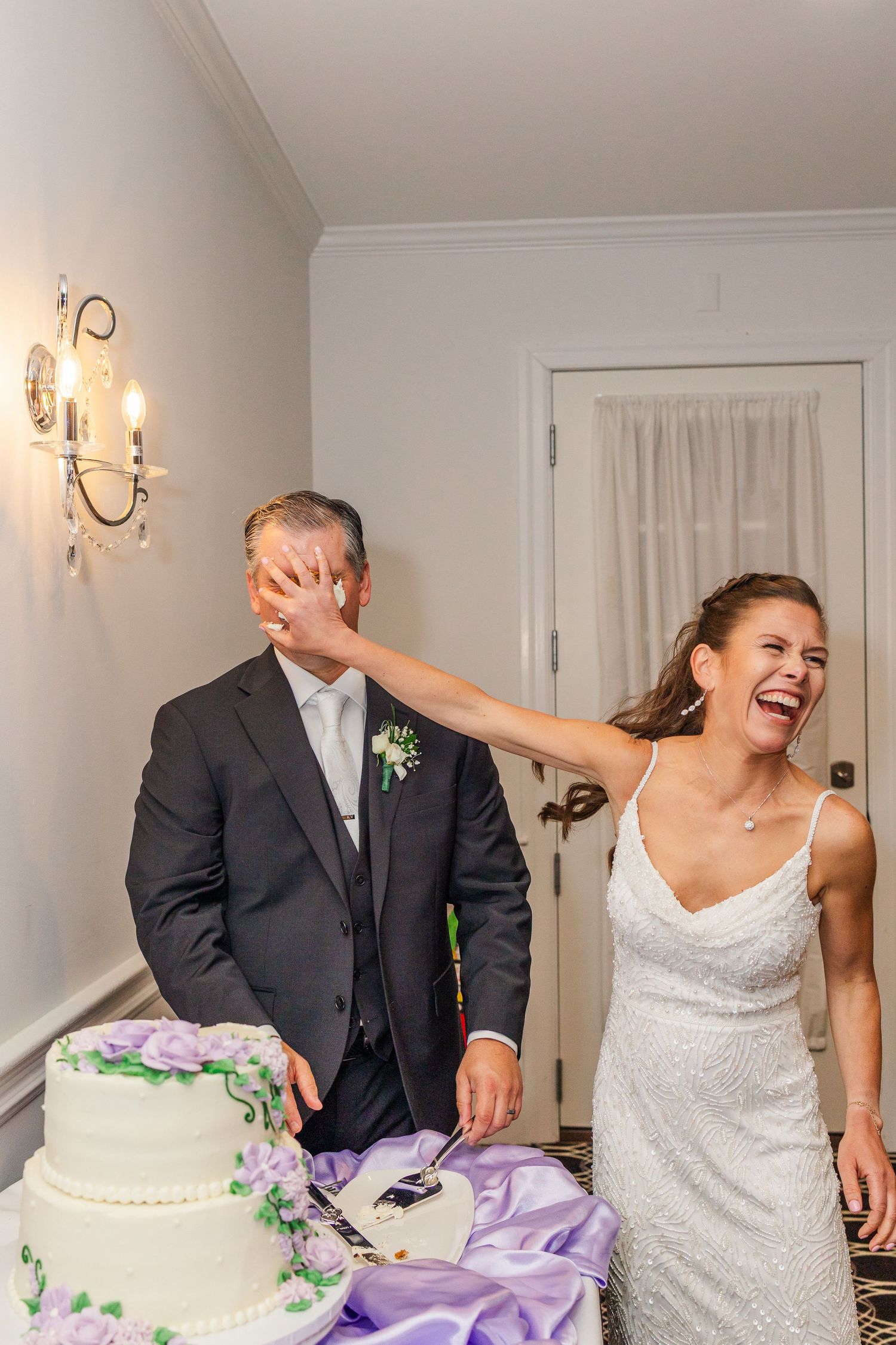 Bride laughing as she smashes wedding cake into groom's face at elegant reception with tiered white cake.