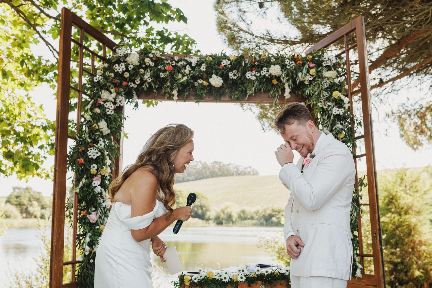 A joyful couple shares a candid moment during their outdoor wedding ceremony under a floral arch by a lake.