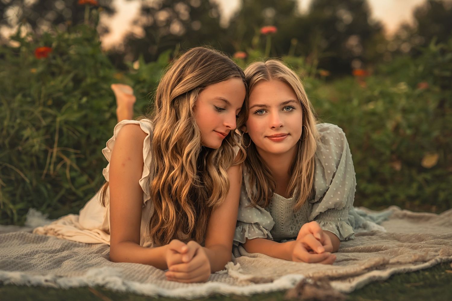 Two young girls with long hair lying on a blanket in a garden, smiling warmly during golden hour.