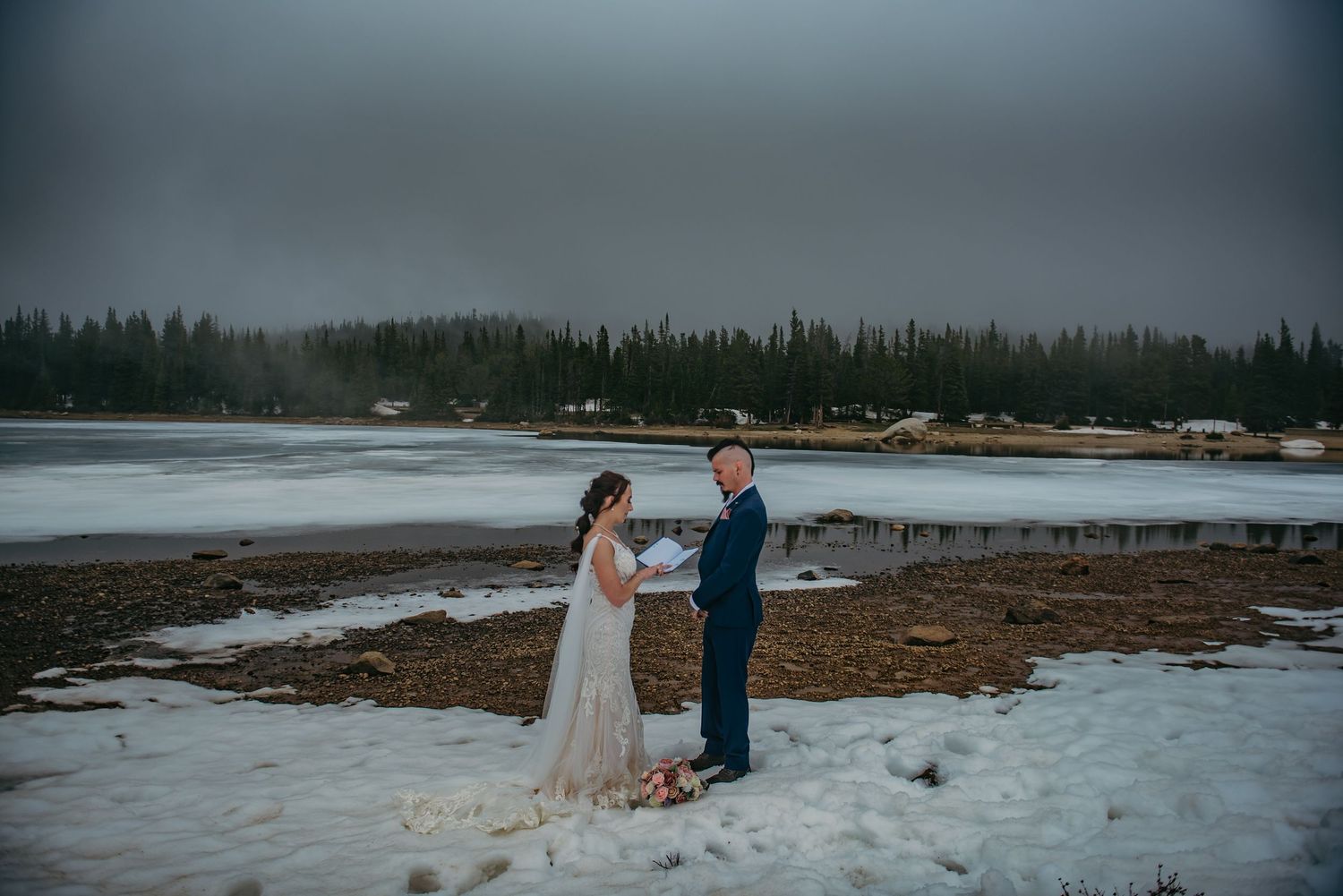Couple exchanging vows outdoors in snowy landscape near frozen lake with evergreen forest backdrop.