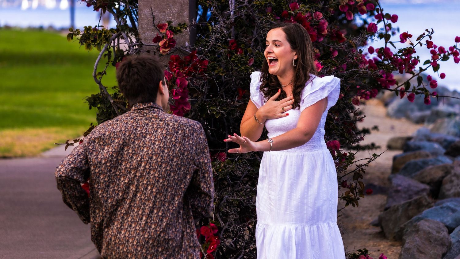 A woman in a white dress reacts expressively to a man in a floral shirt near red flowering bushes outdoors.