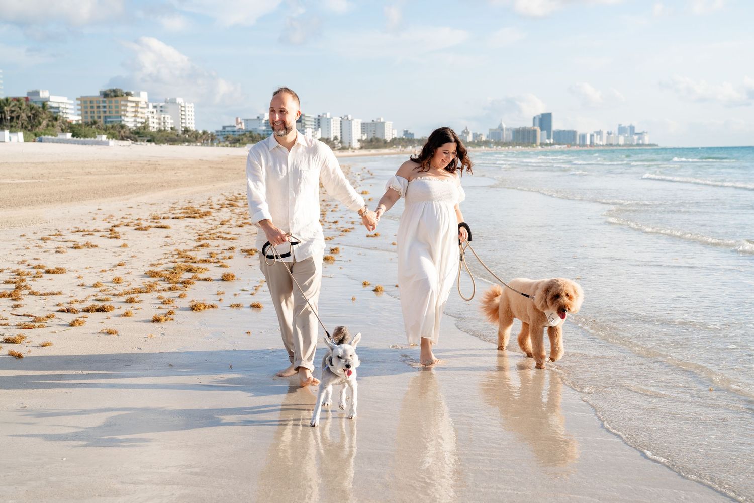 Couple in white outfits walking two dogs on a sunny Miami beach with city skyline in background.