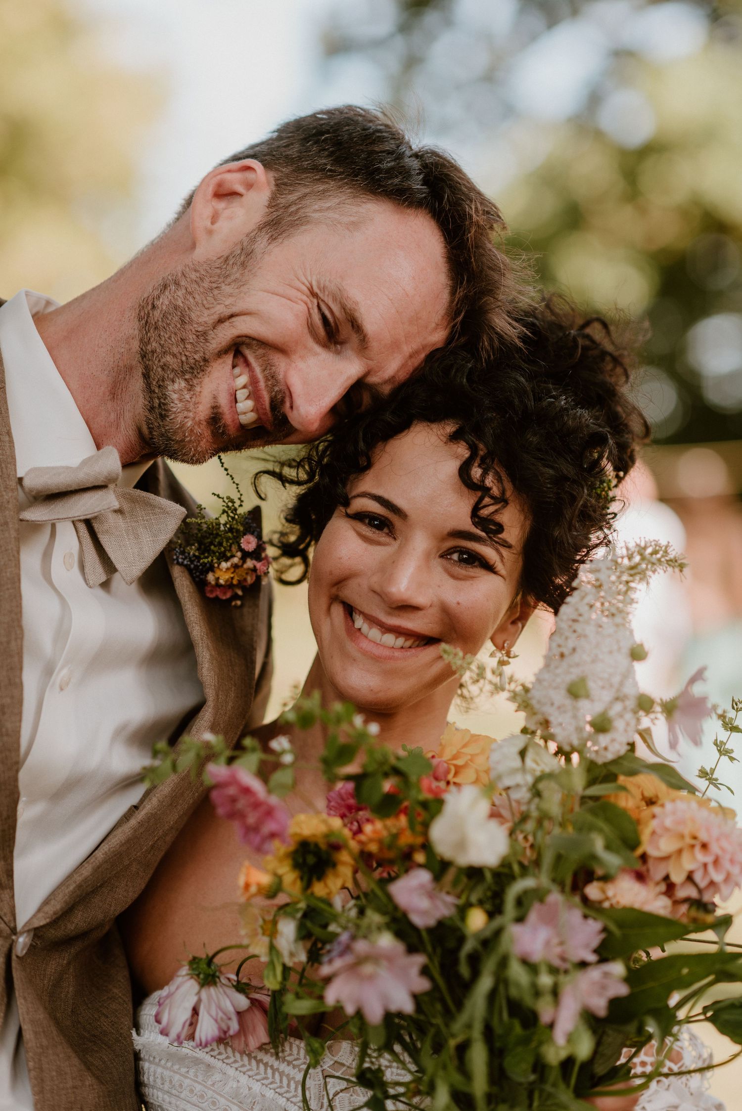 Smiling bride and groom pose closely together with a colorful wildflower bouquet at an outdoor wedding ceremony.