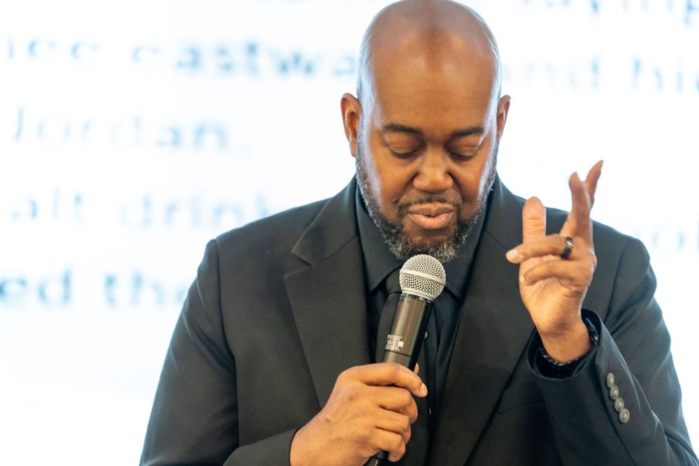 A man in a black suit speaks into a microphone while gesturing with his fingers during a presentation.