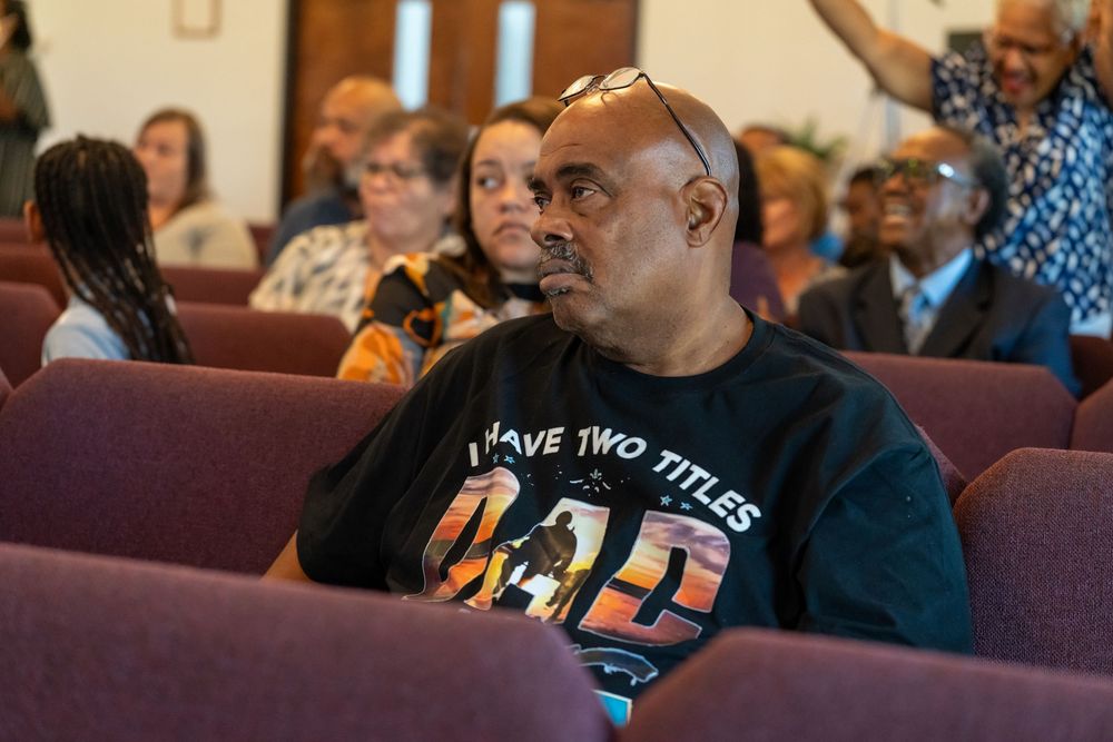 Man in 'I Have Two Titles Dad' shirt sits in maroon church pew, surrounded by diverse congregation members.