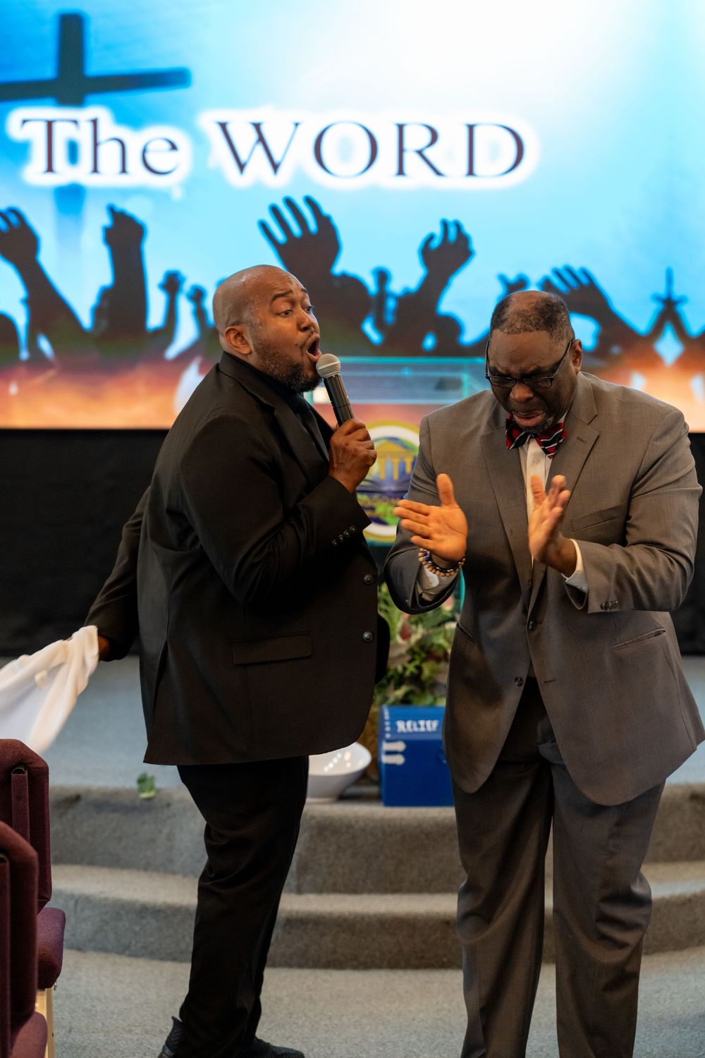 Two men in suits performing worship at a church service with 'The WORD' displayed on screen behind them.