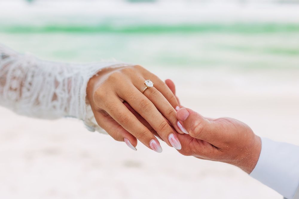 Close-up of a diamond engagement ring on a bride's hand held by groom on a tropical beach.