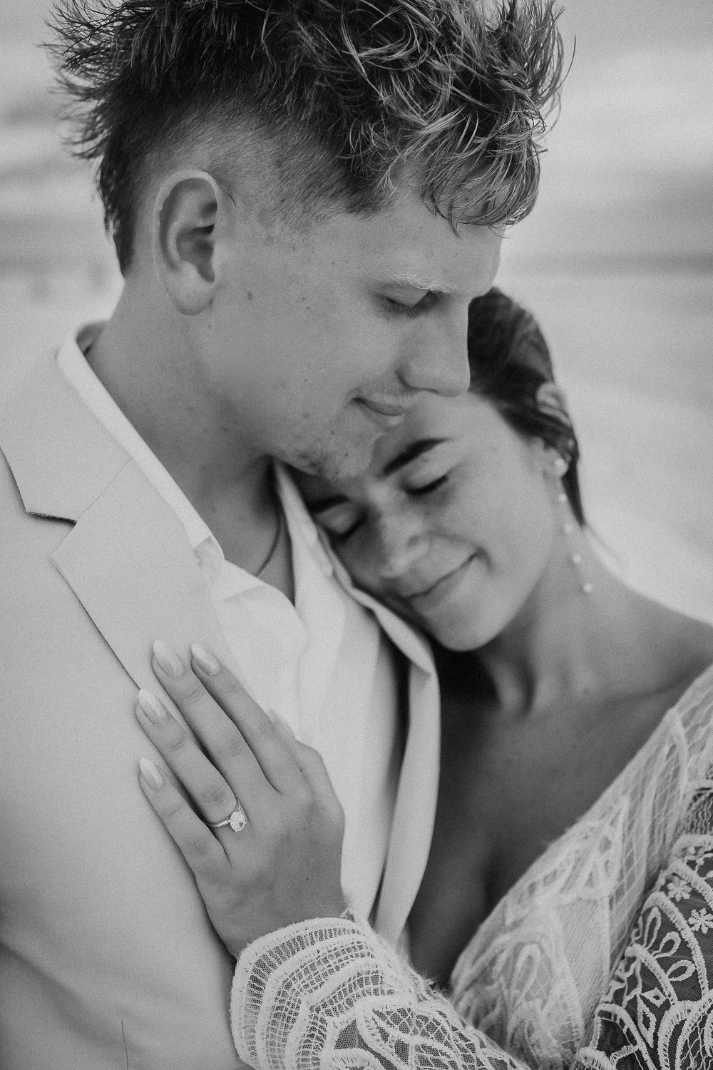 Black and white photo of a couple embracing on a beach, bride in lace dress resting her head on groom's chest.