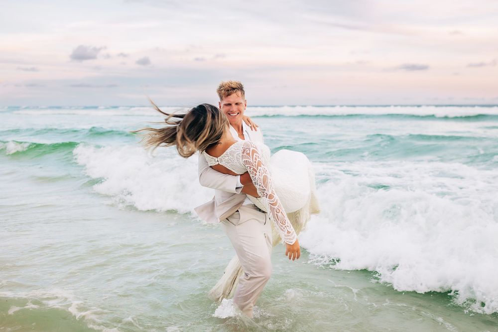 Couple in wedding attire joyfully embracing in ocean waves on a tropical beach at sunset.