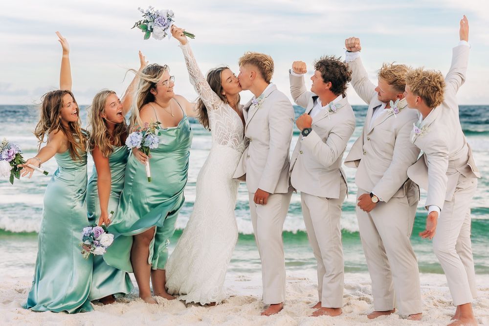 Wedding party celebrating on a beach, bride and groom kissing while bridesmaids in teal dresses and groomsmen in tan suits cheer.