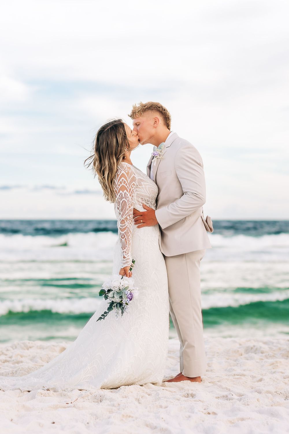 Bride and groom kissing on a white sand beach, bride in lace gown holding bouquet, groom in tan suit, ocean backdrop.