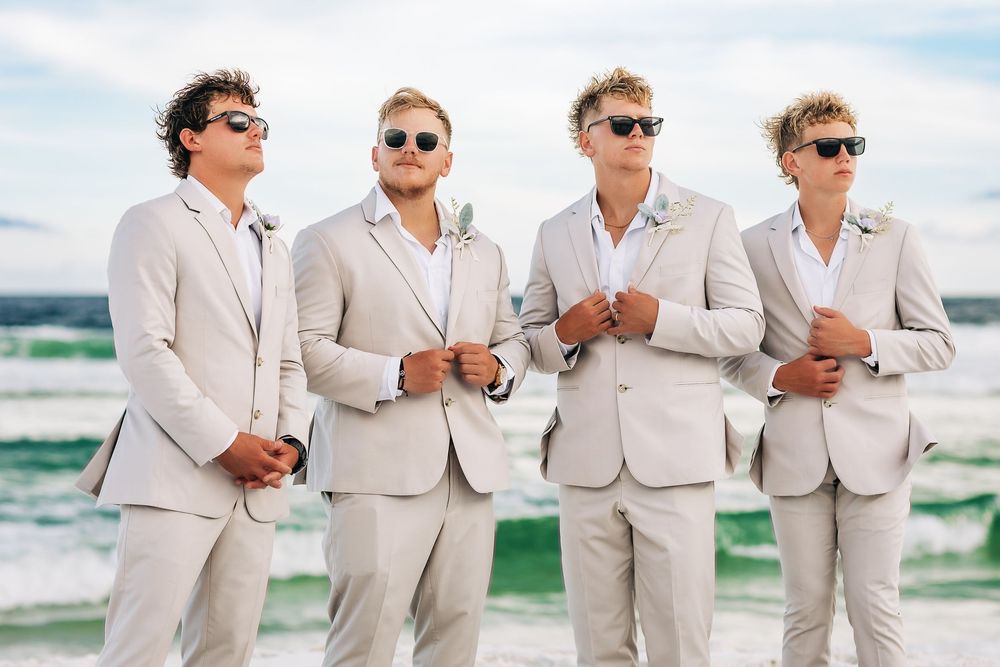 Four groomsmen in matching tan suits and sunglasses pose on a beach with turquoise waves behind them.