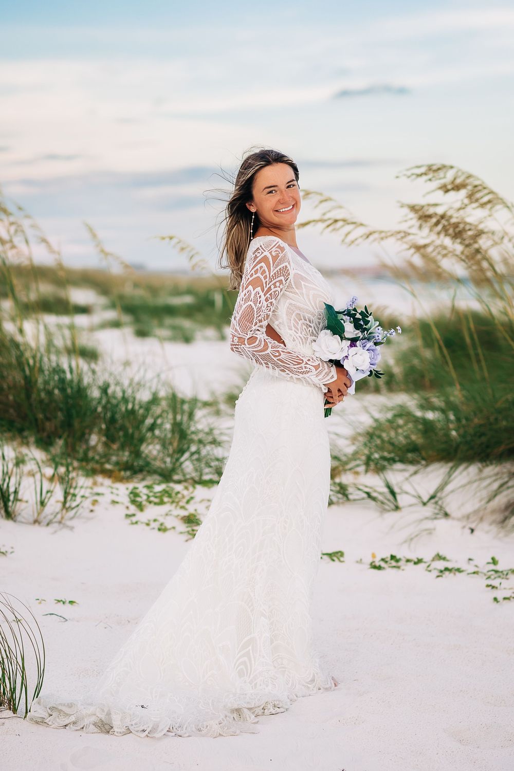 Smiling bride in lace long-sleeve wedding gown holding floral bouquet on white sand beach dunes.