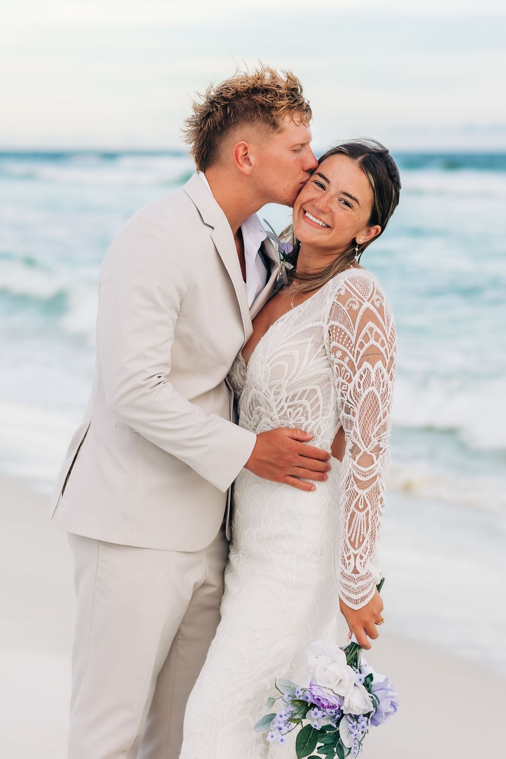 Groom in tan suit kissing bride on cheek at beach wedding, bride smiling holding purple and white bouquet.