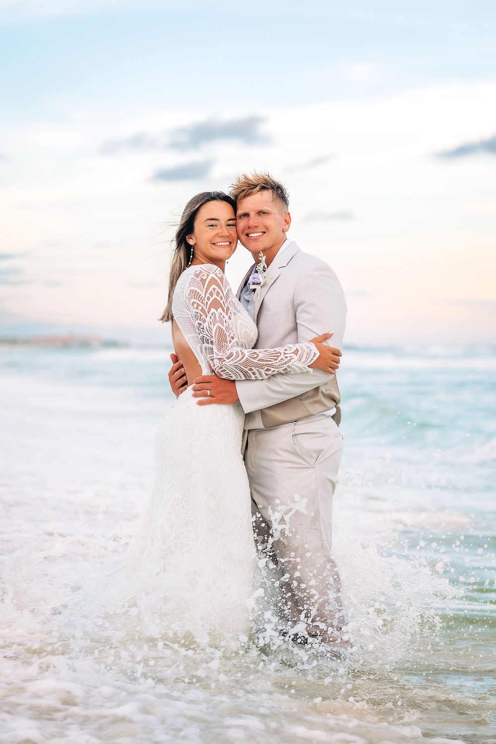 Happy newlywed couple embracing in ocean waves at sunset beach wedding, bride in lace gown and groom in tan suit.