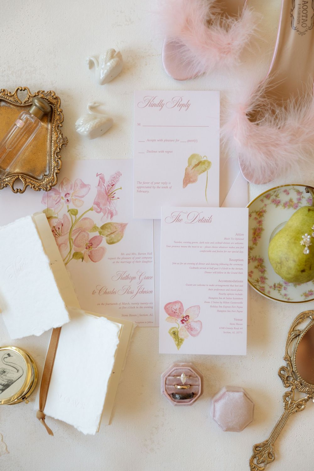 Pink floral wedding invitation suite with RSVP card, details card, ring box, perfume bottle, and feathered heels on white surface.