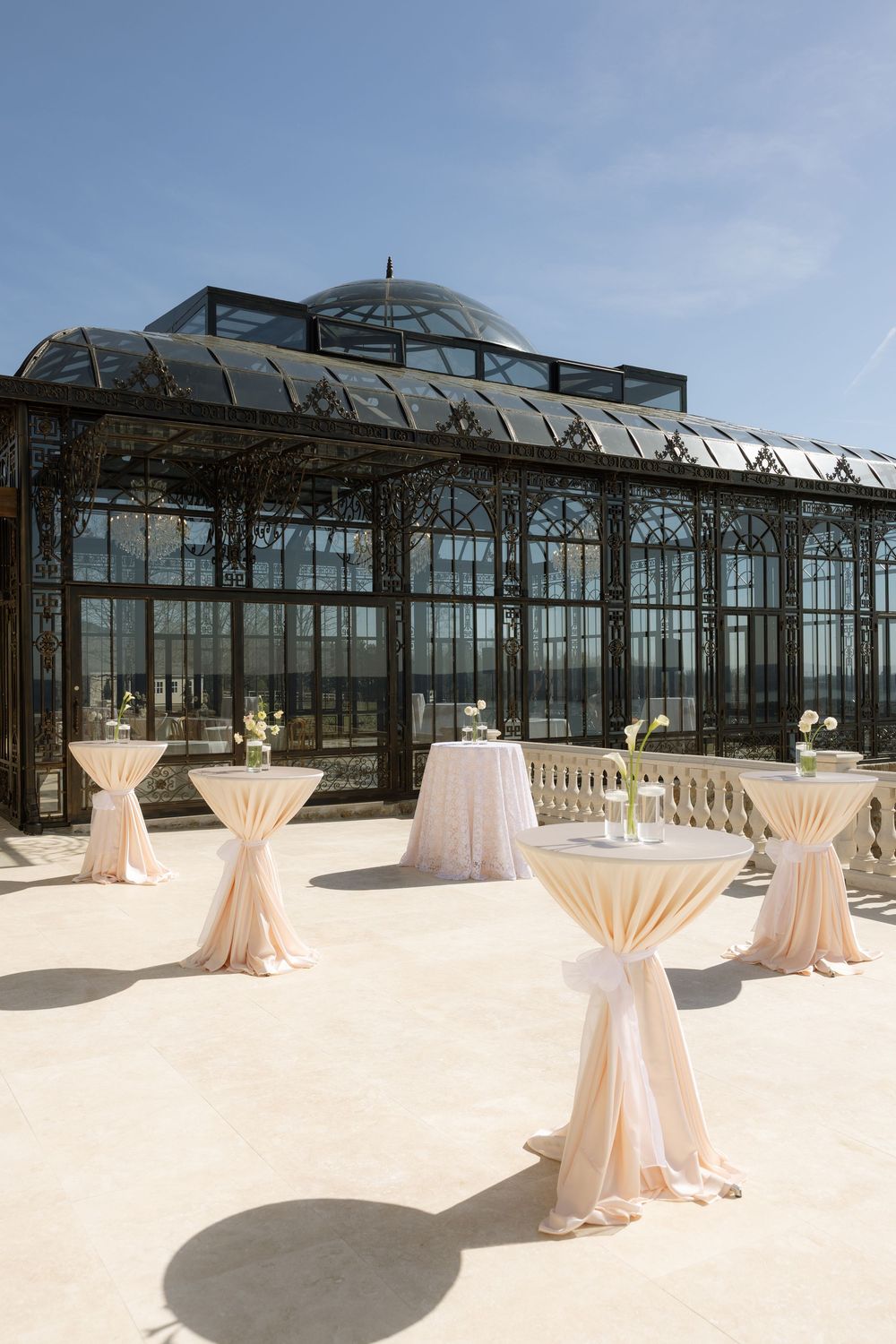 Elegant outdoor cocktail reception with draped bistro tables on a terrace in front of a Victorian glass greenhouse.