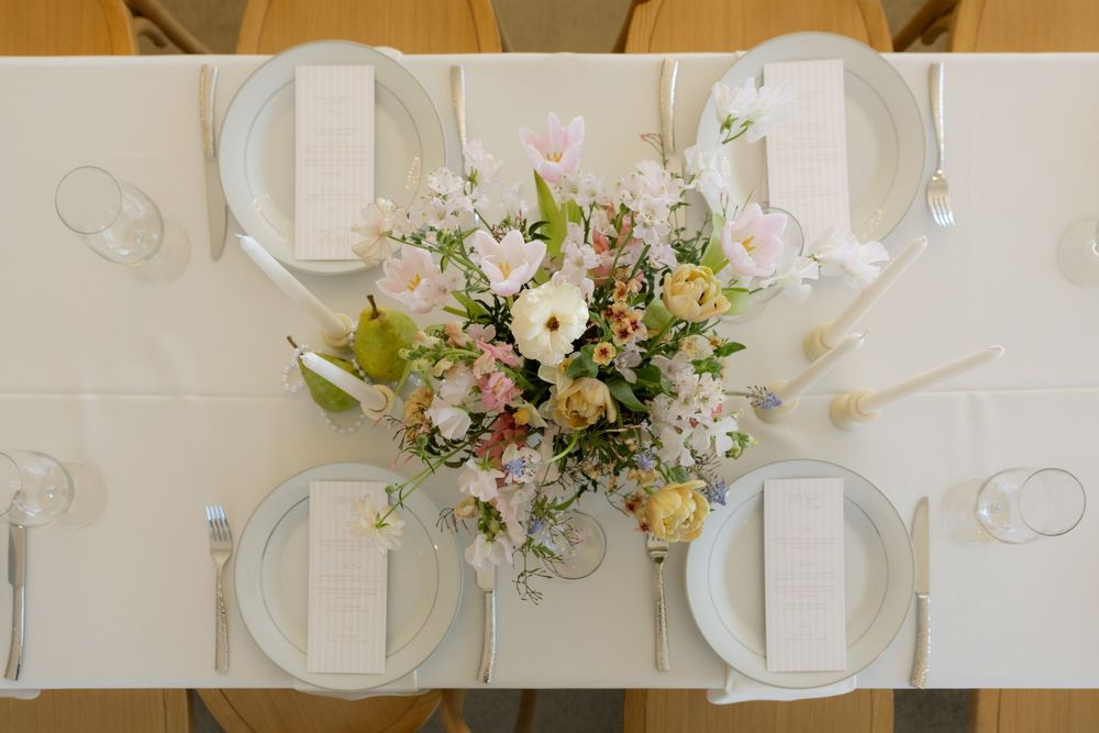 Elegant wedding table setting with pastel floral centerpiece, white linens, gray plates, and menu cards from overhead view.