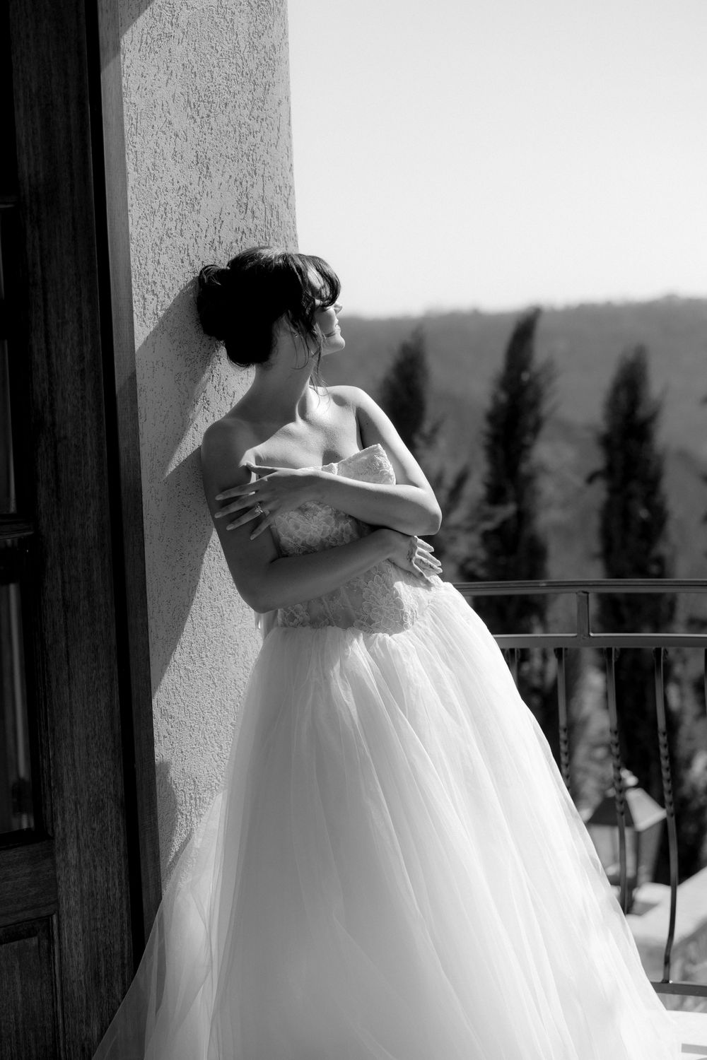 Bride in strapless white ball gown leans against wall on sunlit balcony, arms crossed, in elegant black and white photo.