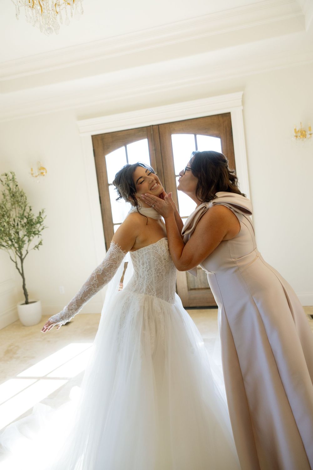 Bride in strapless lace wedding gown shares tender moment with woman in elegant venue before ceremony.