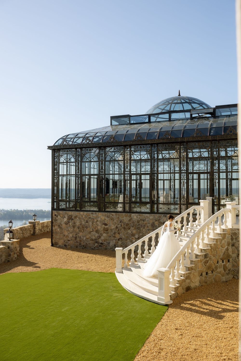 Elegant Victorian greenhouse with glass dome roof, white balustrade staircase, and manicured lawn overlooking a scenic waterfront.