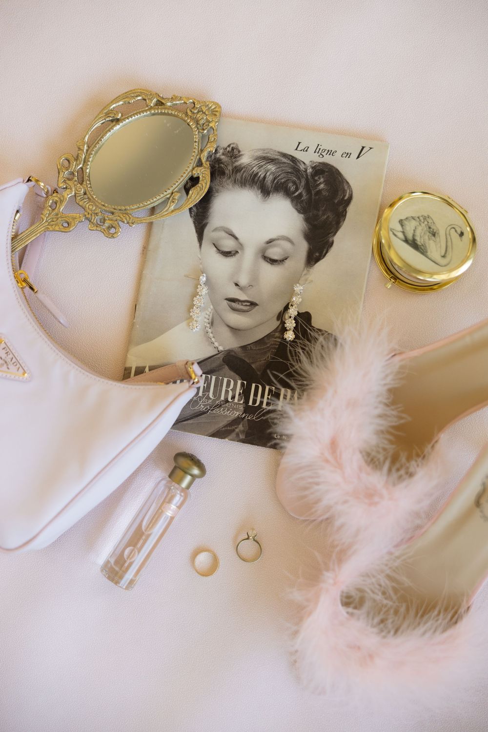 Vintage bridal flat lay with gold hand mirror, French magazine, pink feather mules, perfume, rings, and compact on white background.