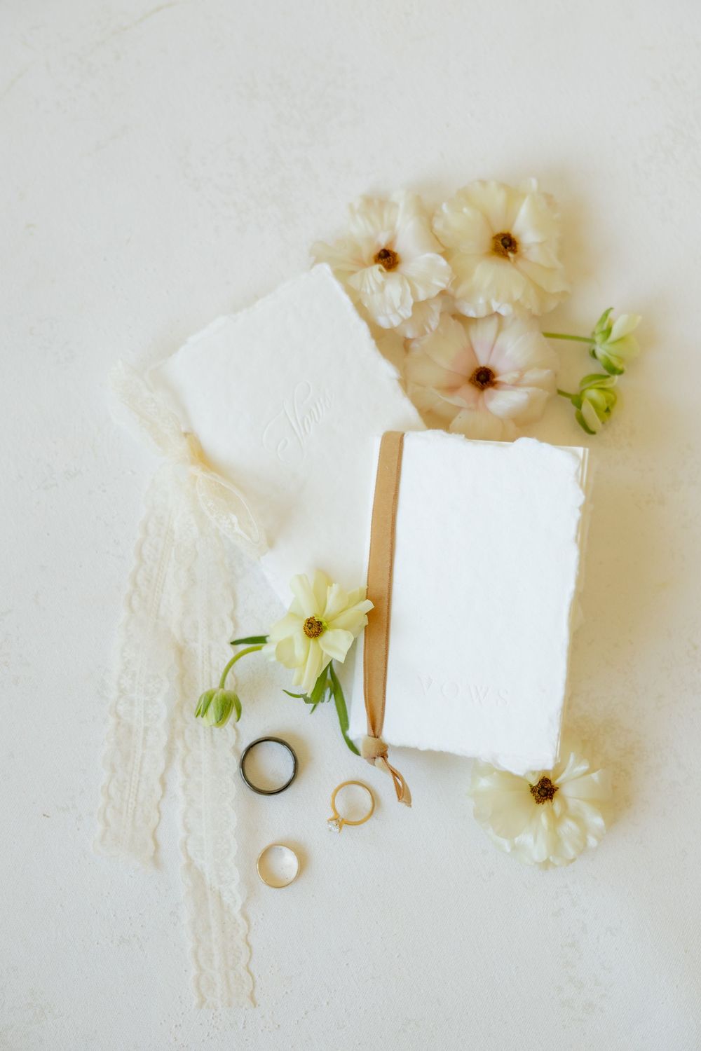 Minimalist wedding flat lay with white deckle edge cards, cream flowers, lace ribbon, and gold and black rings on white surface.