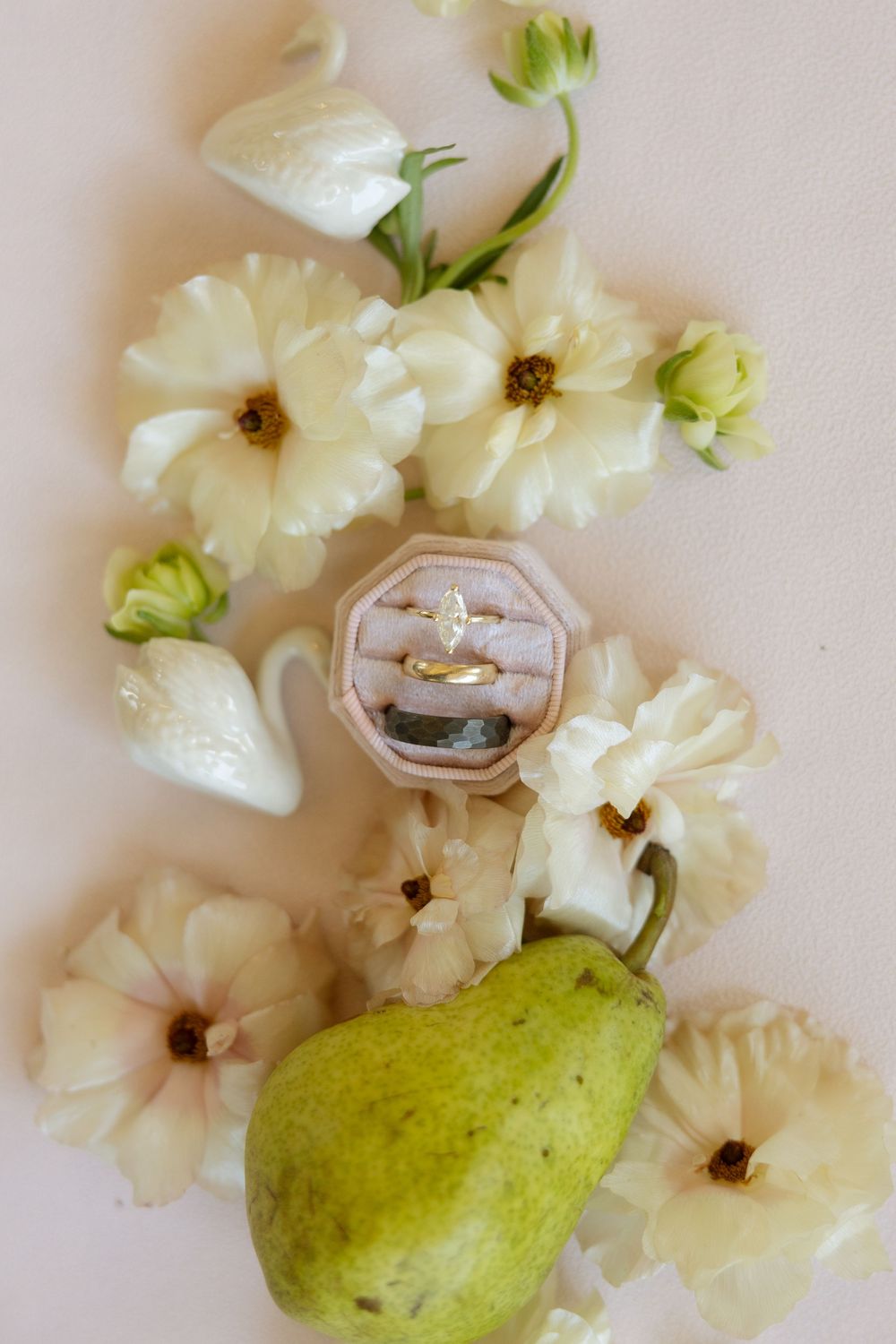Gold engagement ring in velvet box surrounded by white flowers and a green pear on a soft pink background.