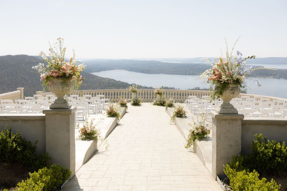 Elegant outdoor wedding ceremony aisle with floral urns overlooking a scenic lake and rolling hills.