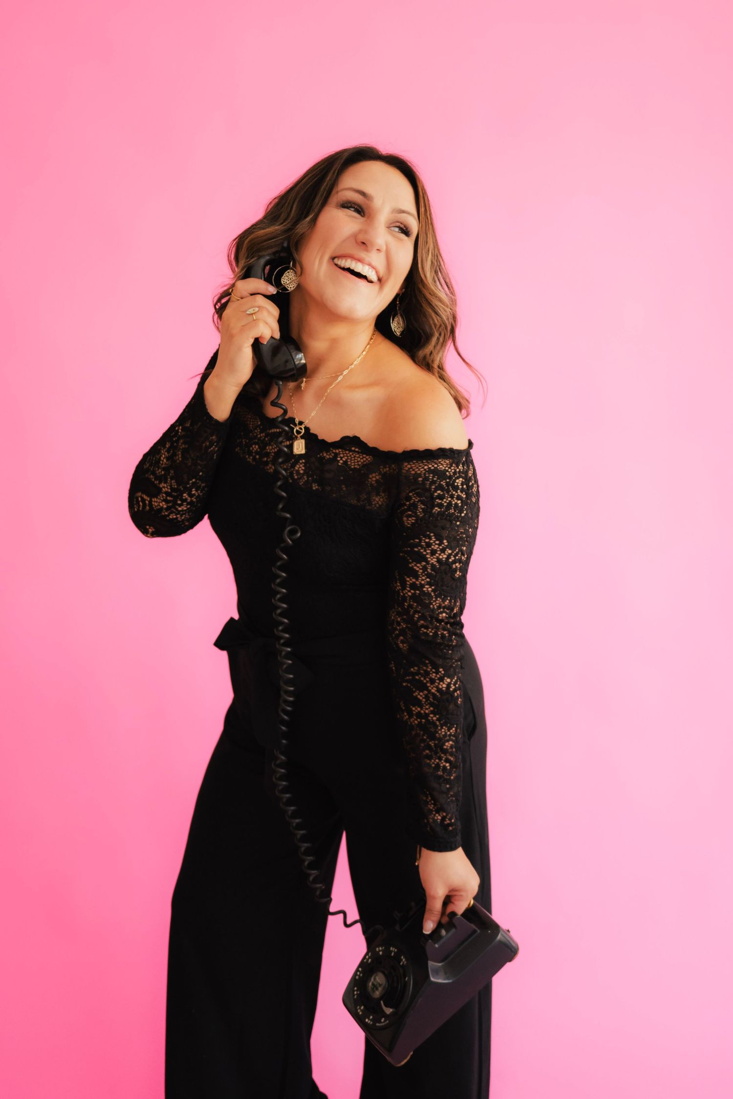 Woman in black lace off-shoulder jumpsuit laughing while holding vintage phone against pink background.
